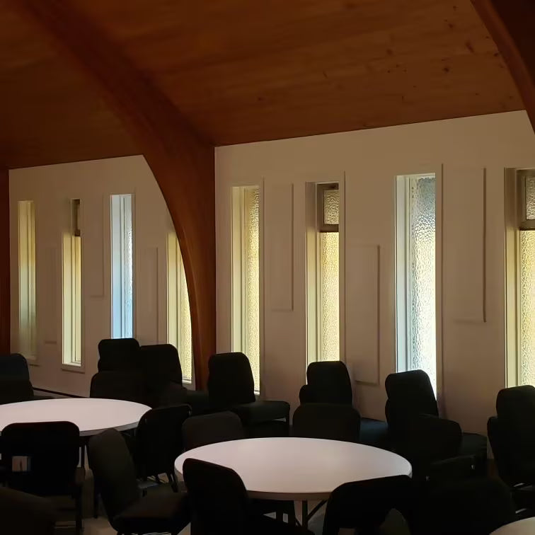 Church fellowship hall with round tables and chairs, showing tall narrow windows and wood beam ceiling