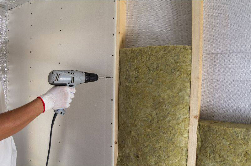 Contractor screwing drywall over mineral wool insulation batts in wall cavity during soundproofing installation