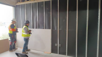 Two construction workers installing soundproofing panels on metal stud wall framing in a commercial space