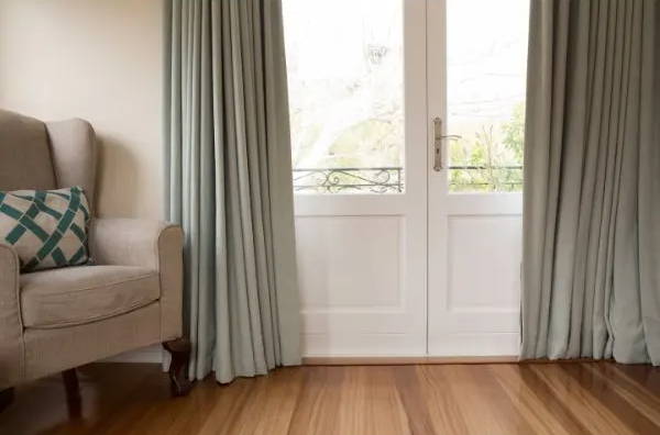 Living room with french doors and sage green curtains beside a beige armchair on hardwood floors
