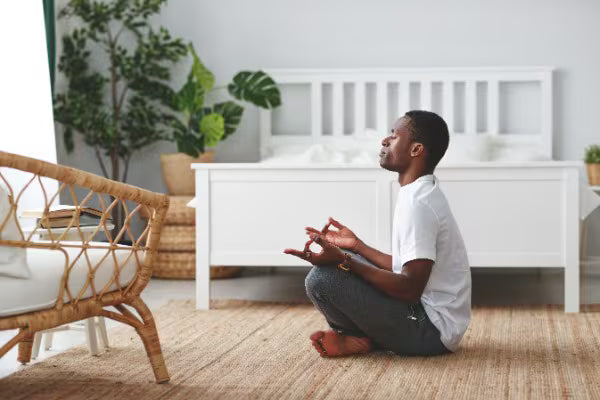 Man meditating in a bedroom with a white bed and wicker chair.